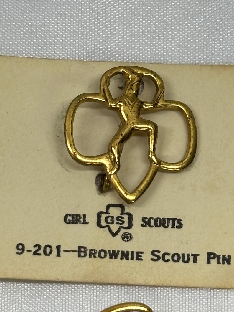 May include: A gold-colored Brownie Scout pin featuring a stylized figure within a trefoil shape. The pin is attached to a tan card with the Girl Scouts logo and the text "9-201-Brownie Scout Pin".