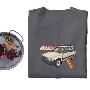 May include: A dark gray sweatshirt featuring a beige 4WD station wagon graphic with a roof rack and the text "4WD". The graphic includes red, orange, and yellow stripes. The sweatshirt is a crew neck style.