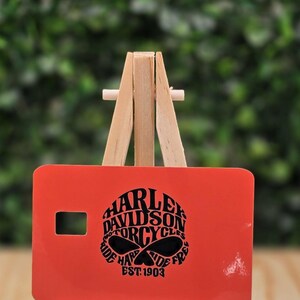 Pu&ograve; includere: Una carta rettangolare arancione con una grafica nera di un teschio e il testo Harley Davidson Motorcycles Ride Hard Ride Free Est. 1903. La carta &egrave; esposta su un piccolo cavalletto di legno, con un ritaglio rettangolare sul lato sinistro.
