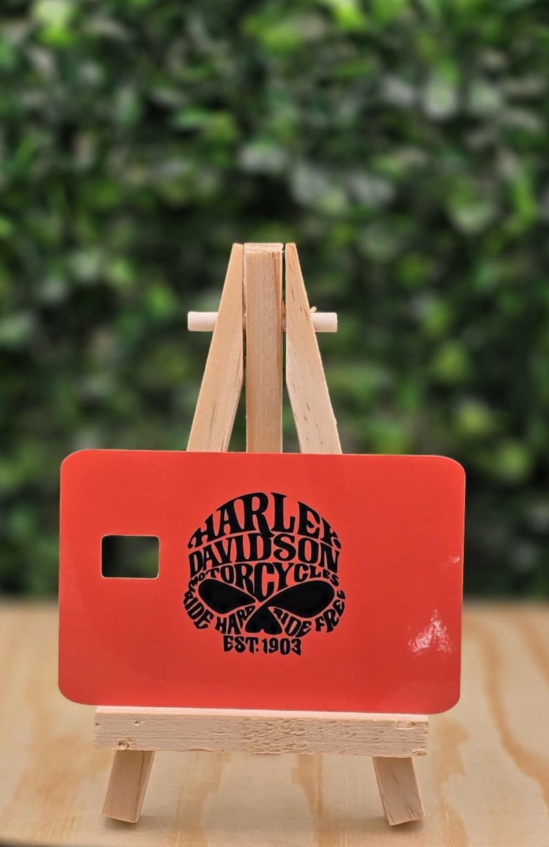Pu&ograve; includere: Una carta rettangolare arancione con una grafica nera di un teschio e il testo Harley Davidson Motorcycles Ride Hard Ride Free Est. 1903. La carta &egrave; esposta su un piccolo cavalletto di legno, con un ritaglio rettangolare sul lato sinistro.