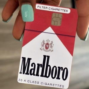 May include: A rectangular credit card skin featuring a red and white design modeled after a pack of Marlboro cigarettes. The card displays the text FILTER CIGARETTES, the Marlboro logo, and the text 20 A CLASS CIGARETTES with a gold EMV chip.