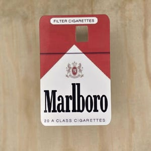 May include: A rectangular Marlboro cigarette pack featuring a red and white design. The top section is red with the text FILTER CIGARETTES. The white lower section displays the Marlboro logo, a crest, and the text 20 A CLASS CIGARETTES.