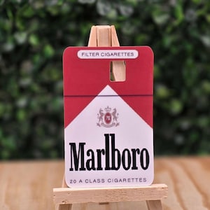 May include: A rectangular card featuring the red and white Marlboro cigarette pack design, with the text FILTER CIGARETTES, the Marlboro logo, and 20 A CLASS CIGARETTES, displayed on a small, light-colored wooden easel stand.