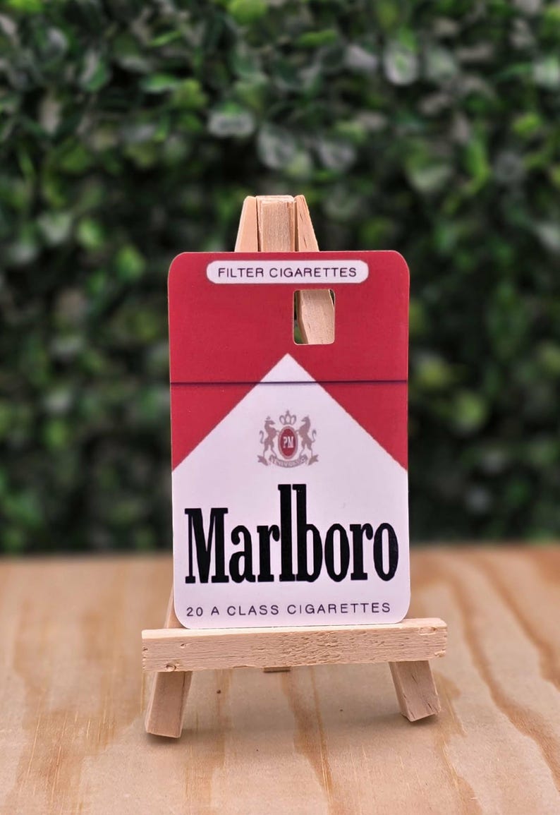 May include: A rectangular card featuring the red and white Marlboro cigarette pack design, with the text FILTER CIGARETTES, the Marlboro logo, and 20 A CLASS CIGARETTES, displayed on a small, light-colored wooden easel stand.