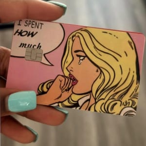 May include: A rectangular credit card featuring a pop art illustration of a blonde person with a tear on their cheek. A speech bubble contains the text I SPENT HOW much. The card has a pink background and a metallic EMV chip embedded on the left side.