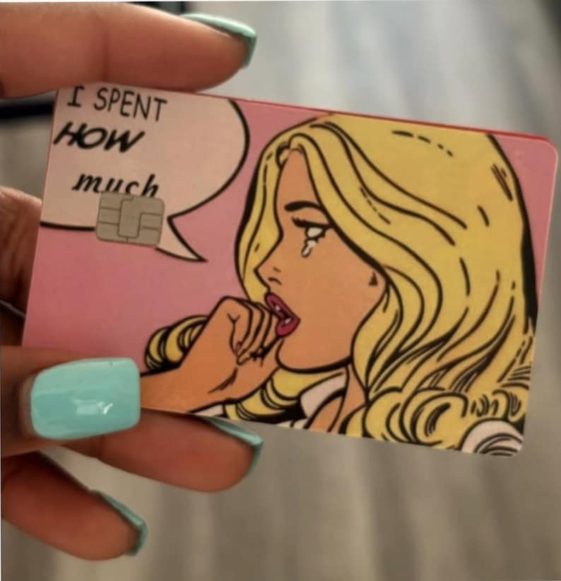 May include: A rectangular credit card featuring a pop art illustration of a blonde person with a tear on their cheek. A speech bubble contains the text I SPENT HOW much. The card has a pink background and a metallic EMV chip embedded on the left side.