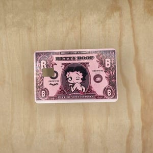 May include: A rectangular pink sticker designed to look like a banknote featuring Betty Boop. The text reads Boop Oop A Doop, Betty Boop, 08091930, and Five Hundred Kisses. It includes a square cutout on the left side and the letter B in corners.