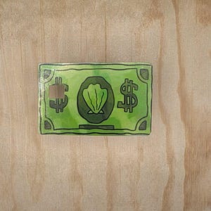 May include: A rectangular green pin featuring a central dark green circle with a light green seashell illustration. The design includes black dollar signs on the left and right sides, a small square cutout on the left, and dark green corner accents.