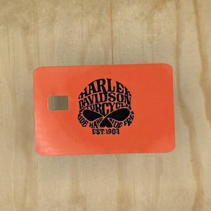 Pu&ograve; includere: Un adesivo rettangolare arancione per carta di credito con una grafica nera a forma di teschio composta dal testo Harley Davidson Motorcycles Ride Hard Ride Free Est. 1903. L'adesivo presenta un piccolo ritaglio rettangolare per il chip.