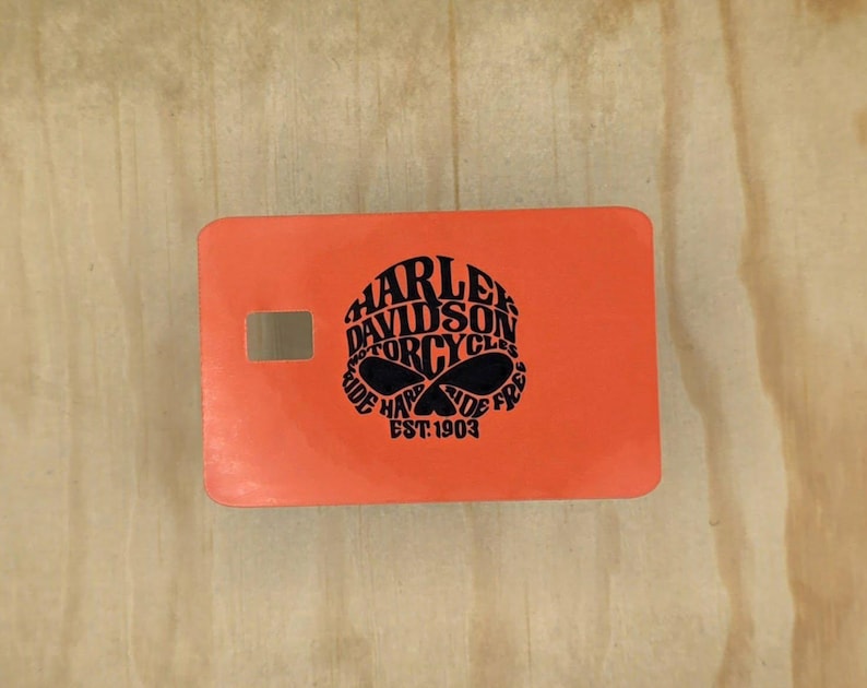 Pu&ograve; includere: Un adesivo rettangolare arancione per carta di credito con una grafica nera a forma di teschio composta dal testo Harley Davidson Motorcycles Ride Hard Ride Free Est. 1903. L'adesivo presenta un piccolo ritaglio rettangolare per il chip.