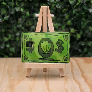 May include: A rectangular green card featuring a central illustration of a seashell inside a circle, flanked by two dollar sign symbols, displayed on a small wooden tripod easel. The card has a small square cutout on the left side.