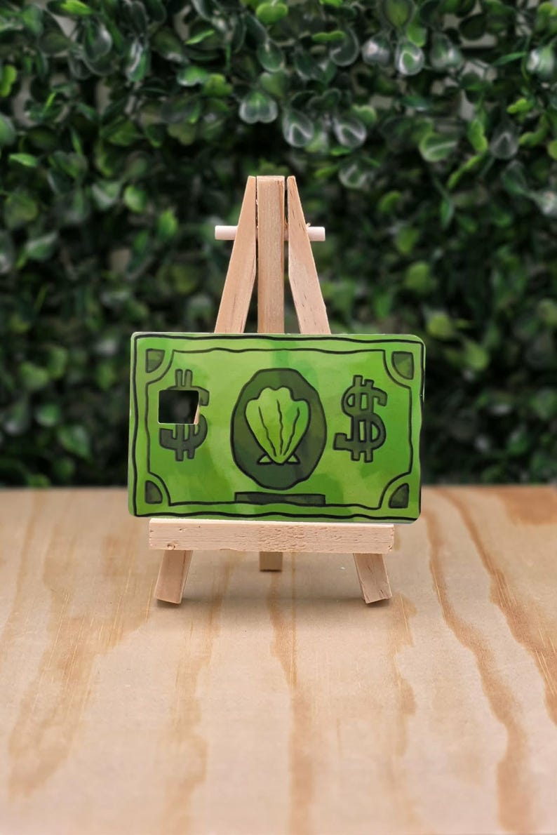 May include: A rectangular green card featuring a central illustration of a seashell inside a circle, flanked by two dollar sign symbols, displayed on a small wooden tripod easel. The card has a small square cutout on the left side.