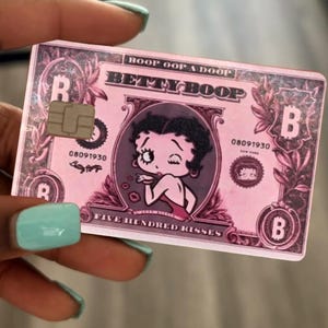 May include: A rectangular pink bank card featuring a central illustration of Betty Boop winking. The card includes the text BOOP OOP A DOOP, BETTY BOOP, 08091930, NEW YORK, and FIVE HUNDRED KISSES, along with a metallic chip and Bitcoin symbols.
