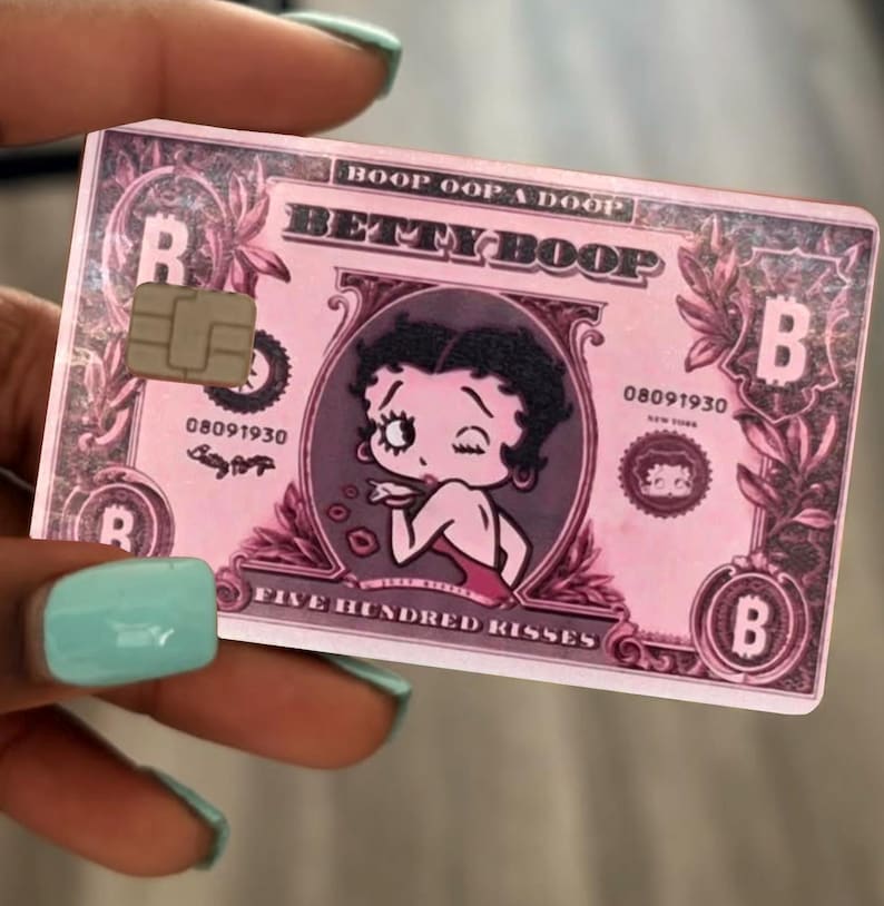 May include: A rectangular pink bank card featuring a central illustration of Betty Boop winking. The card includes the text BOOP OOP A DOOP, BETTY BOOP, 08091930, NEW YORK, and FIVE HUNDRED KISSES, along with a metallic chip and Bitcoin symbols.