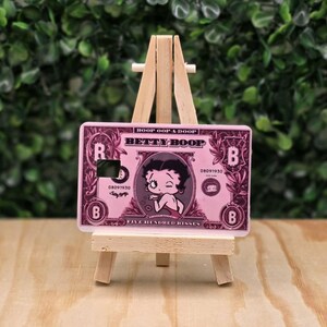 May include: A rectangular pink credit card skin featuring a Betty Boop illustration, the text BOOP OOP A DOOP, BETTY BOOP, and FIVE HUNDRED KISSES, with a square cutout, displayed on a small wooden easel stand for decorative display purposes.