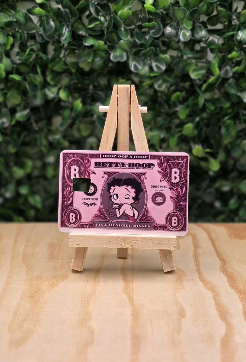 May include: A rectangular pink credit card skin featuring a Betty Boop illustration, the text BOOP OOP A DOOP, BETTY BOOP, and FIVE HUNDRED KISSES, with a square cutout, displayed on a small wooden easel stand for decorative display purposes.