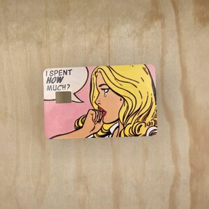 May include: A rectangular credit card skin featuring a pop art illustration of a blonde person with a tear on their cheek. The text I SPENT HOW MUCH? appears in a white speech bubble against a pink background. The card has a rectangular cutout.