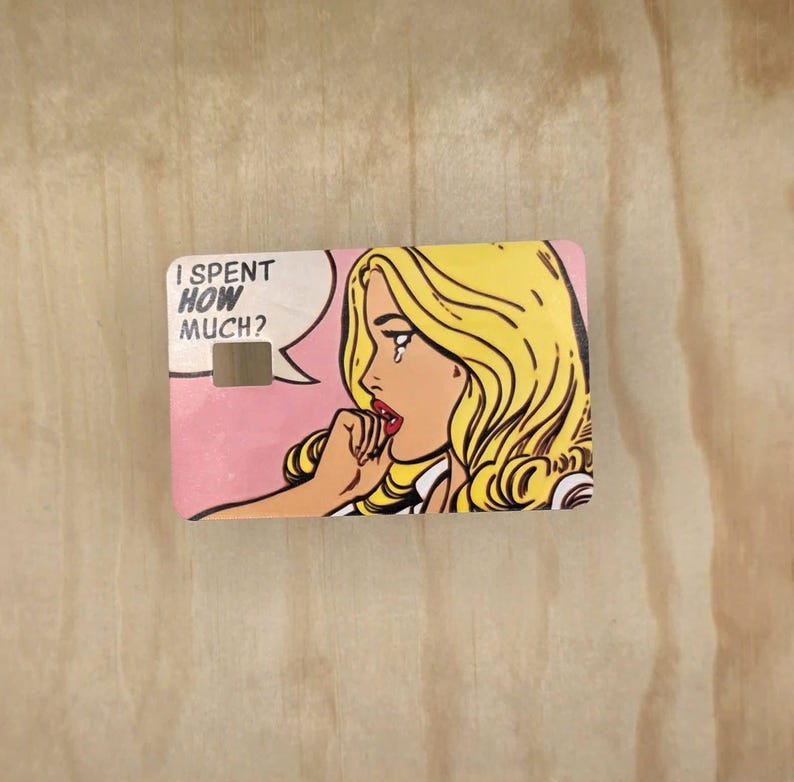 May include: A rectangular credit card skin featuring a pop art illustration of a blonde person with a tear on their cheek. The text I SPENT HOW MUCH? appears in a white speech bubble against a pink background. The card has a rectangular cutout.
