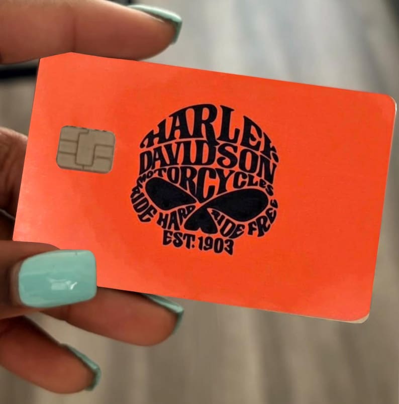 Pu&ograve; includere: Una carta di credito rettangolare color arancione brillante con un logo grafico nero al centro. Il logo contiene il testo Harley Davidson Motorcycles, Ride Hard Ride Free e Est. 1903. Un chip EMV metallico argentato si trova a sinistra.
