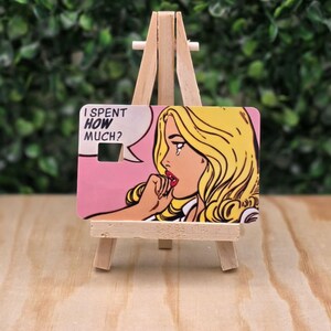 May include: A rectangular plastic card featuring a pop art illustration of a blonde person with a tear on their cheek, hand to their mouth, and a speech bubble containing the text I SPENT HOW MUCH? The card has a small square cutout.