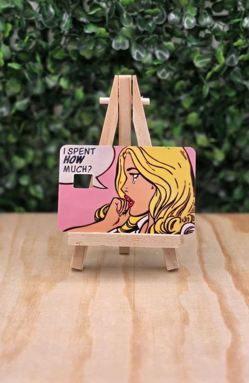 May include: A rectangular plastic card featuring a pop art illustration of a blonde person with a tear on their cheek, hand to their mouth, and a speech bubble containing the text I SPENT HOW MUCH? The card has a small square cutout.