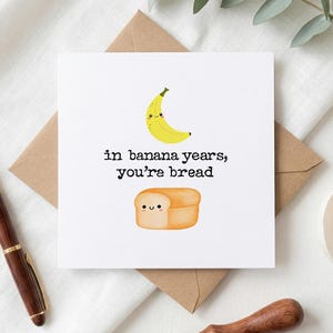 May include: A white greeting card with the text "in banana years, you're bread" and illustrations of a smiling banana and a loaf of bread. The card is on a brown envelope, with a pen and other items in the background.