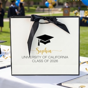 May include: A white graduation gift bag with a black border and a black bow. The bag features a graduation cap graphic and the name "Sophia" in gold script, along with "University of California Class of 2026."