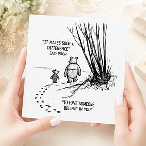 Op de afbeelding: Een vierkante witte wenskaart met een zwart-witte lijntekening van Winnie de Poeh en Knorretje die naar een boom lopen. De tekst luidt: "IT MAKES SUCH A DIFFERENCE" SAID POOH "TO HAVE SOMEONE BELIEVE IN YOU" met voetstappen.