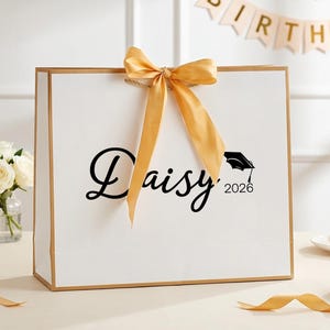 May include: A white gift bag with gold trim and a gold bow. The bag features the name "Daisy" in a script font, along with a graduation cap and the year "2026". The bag is on a table with flowers and desserts.
