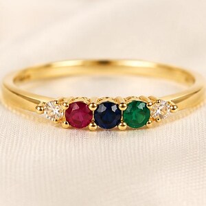 Könnte beinhalten: Ein Goldring mit einer Reihe von fünf rund geschliffenen Edelsteinen. Die mittleren Steine sind ein roter Rubin, ein blauer Saphir und ein grüner Smaragd, flankiert von zwei klaren weißen Steinen auf jeder Seite, gefasst in einem gelben Goldband.