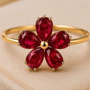 May include: A gold ring featuring a flower-shaped centrepiece composed of five pear-cut, deep red gemstones arranged around a central gold bead. Each gemstone is secured by small gold prongs, and the band is a smooth, polished gold metal.