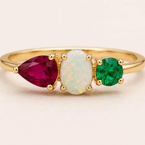 Pode incluir: Um anel de ouro com três pedras cravadas em linha. A pedra central é um opala branco de formato oval com reflexos iridescentes. À esquerda, um rubi vermelho profundo em forma de pera e, à direita, uma esmeralda verde vibrante.
