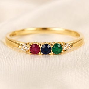 May include: A gold ring featuring five round-cut gemstones set in a row. The center stones include a red ruby, a dark blue sapphire, and a green emerald, flanked by two smaller clear white stones on each side, all held in a polished gold band.