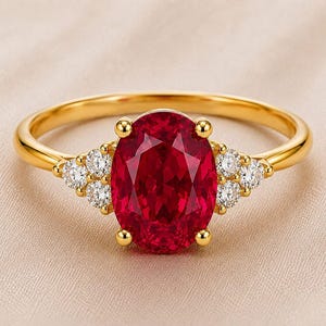 Op de afbeelding: Een gouden ring met een grote, ovaal geslepen, dieprode edelsteen in het midden. De centrale steen wordt aan weerszijden geflankeerd door een groepje van drie kleine, ronde, heldere briljant geslepen stenen. De ringband is van glad, gepolijst geelgoud.
