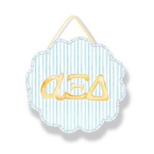 May include: A scalloped, circular wall hanging featuring a light blue and white vertical striped pattern. Centered on the surface are three gold-colored Greek letters, alpha, xi, and delta, rendered in a script style. A yellow cord is attached for hanging.
