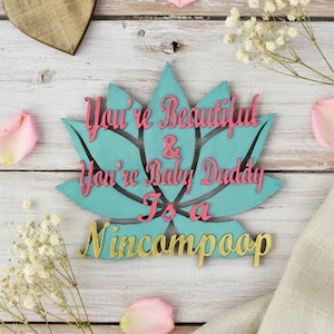 Może przedstawiać: Drewniana dekoracja w kształcie kwiatu lotosu w kolorze turkusowym z napisem "You're Beautiful & You're Baby Daddy Is a Nincampoop" w kolorze różowym i złotym. Zawiera drewniane serce i płatki kwiatów.