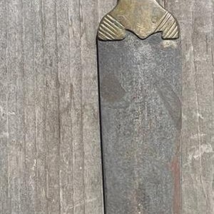 May include: A vintage metal bottle opener with a long, flat, weathered grey handle and a brass-coloured decorative collar featuring etched line details. The tool has a rounded, open-loop head designed for removing bottle caps from glass beverage bottles.