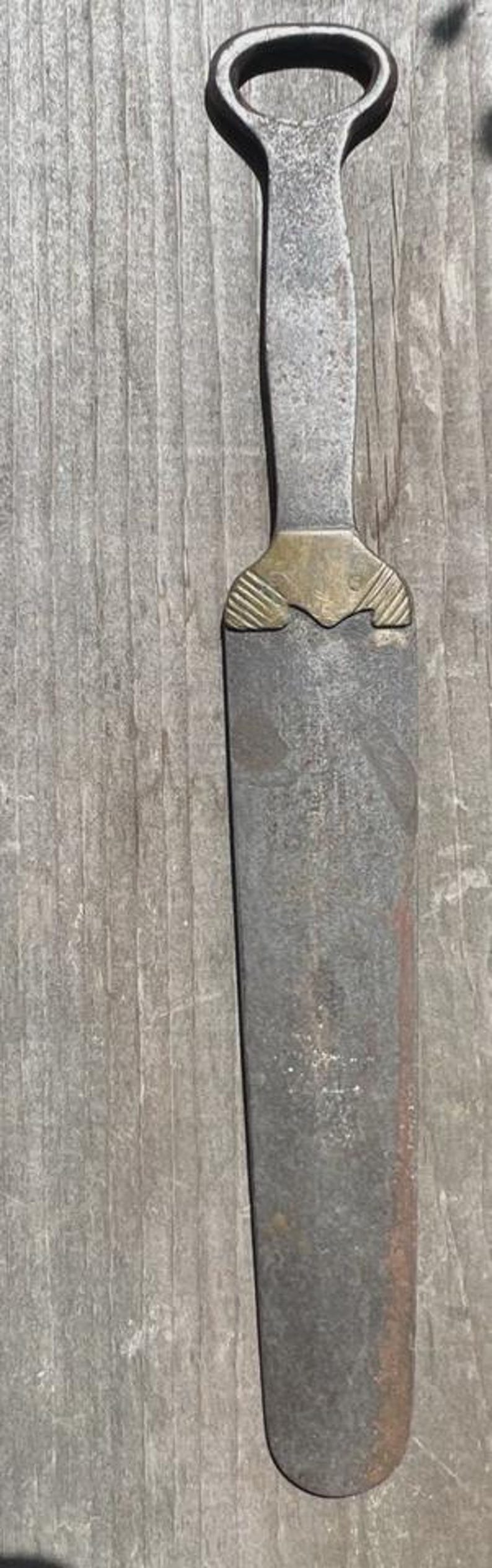 May include: A vintage metal bottle opener with a long, flat, weathered grey handle and a brass-coloured decorative collar featuring etched line details. The tool has a rounded, open-loop head designed for removing bottle caps from glass beverage bottles.
