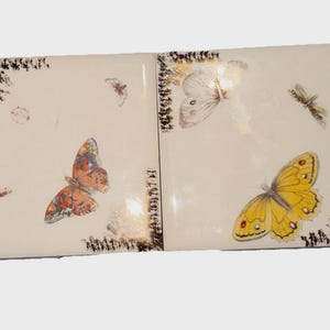 May include: Two white ceramic tiles with butterfly illustrations. One tile features a large orange butterfly and a smaller one. The other tile has a yellow butterfly and a white butterfly. Each tile has a decorative black border.