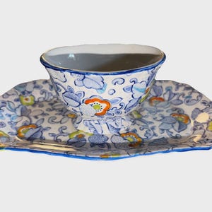 May include: A ceramic teacup and saucer set with a floral pattern in blue, orange, and yellow. The teacup sits on a pedestal base, and the saucer has a scalloped edge. The set is painted with a blue trim.