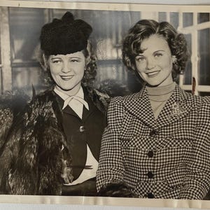 May include: Black and white photograph of two women smiling. One woman wears a dark hat, fur coat, and a dark suit with a white bow tie. The other woman wears a checkered blazer with a brooch.