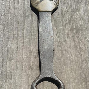 May include: A metal bottle opener featuring a long, weathered iron handle with a dark, textured finish. The neck of the opener is adorned with a decorative brass-toned metal collar that has a pointed, chevron-like shape and etched line details.