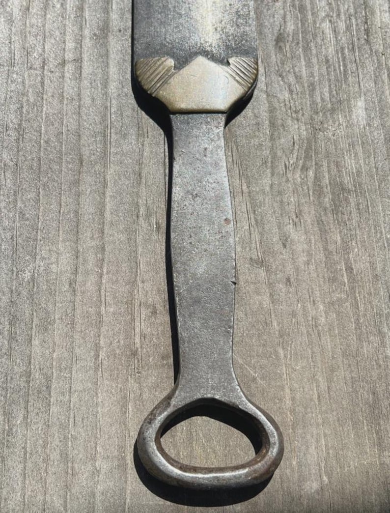 May include: A metal bottle opener featuring a long, weathered iron handle with a dark, textured finish. The neck of the opener is adorned with a decorative brass-toned metal collar that has a pointed, chevron-like shape and etched line details.