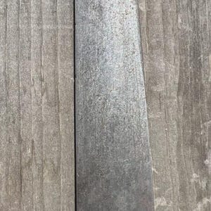 May include: A long, flat, silver-toned metal spatula with a rounded tip and a textured surface. The base of the blade features a decorative brass-coloured metal bolster with a scalloped edge and etched line details, connecting to a handle.