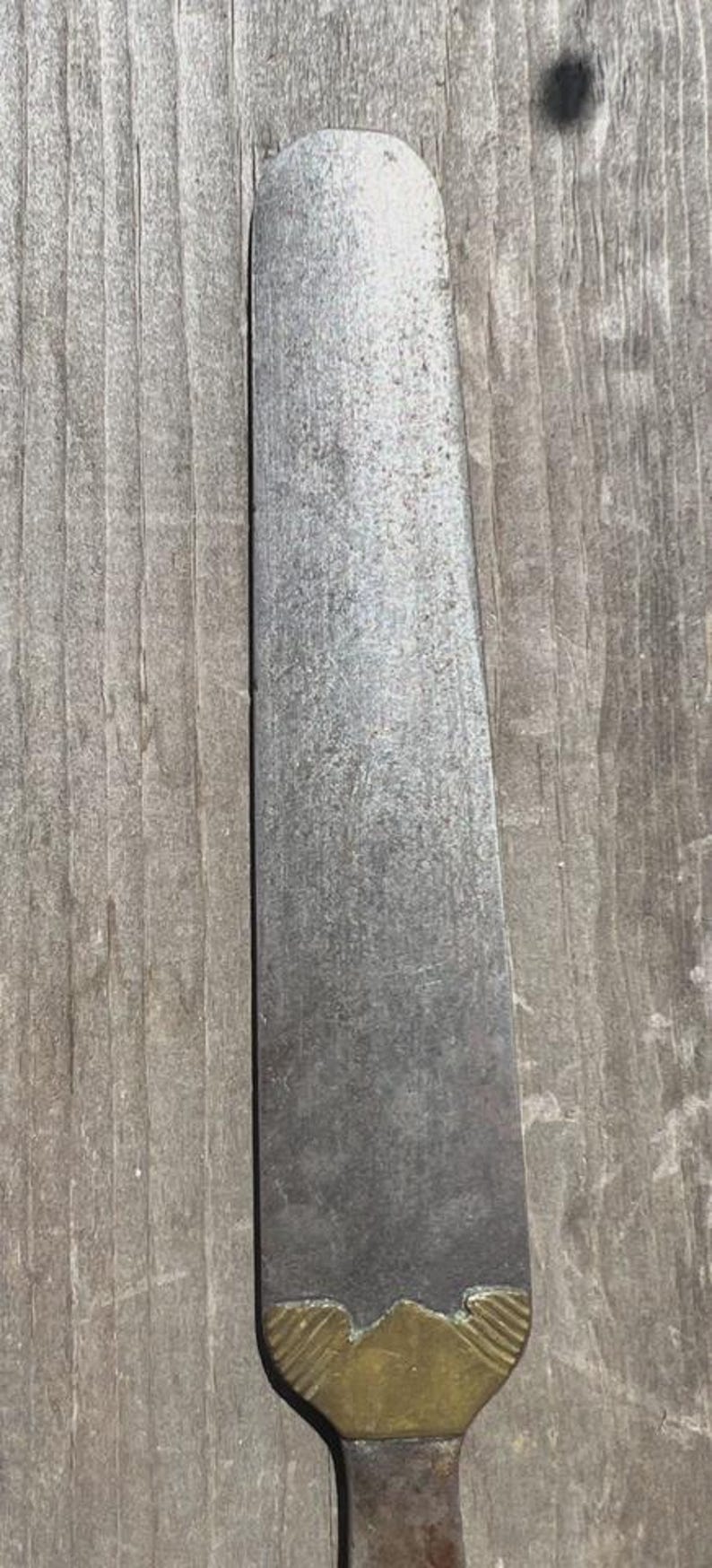 May include: A long, flat, silver-toned metal spatula with a rounded tip and a textured surface. The base of the blade features a decorative brass-coloured metal bolster with a scalloped edge and etched line details, connecting to a handle.