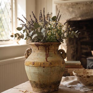 May include: A weathered, rustic ceramic vase with two side handles, featuring a distressed finish in shades of muted green, yellow, and brown. The vase is filled with a bouquet of purple lavender, green eucalyptus leaves, and small white daisies.