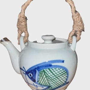 May include: White ceramic teapot with a blue and green fish design. The teapot has a spout, a lid with a small knob, and a rope handle. The handle is made of thick, twisted rope. The fish design is hand-painted.