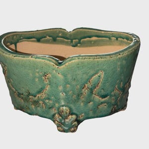 May include: A turquoise ceramic planter with a textured, crackled surface. The planter has a unique, scalloped rim and three decorative feet. The interior is a light tan color, contrasting with the exterior's aged appearance. A decorative piece for plants.