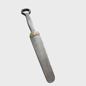 May include: A long, flat, dark grey metal tool with a rounded handle featuring a circular opening at the top. The tool has a smooth, elongated blade section connected to the handle by a decorative, triangular brass-coloured metal accent piece.