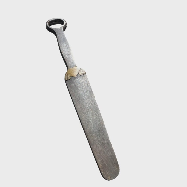 May include: A long, flat, dark grey metal tool with a rounded handle featuring a circular opening at the top. The tool has a smooth, elongated blade section connected to the handle by a decorative, triangular brass-coloured metal accent piece.